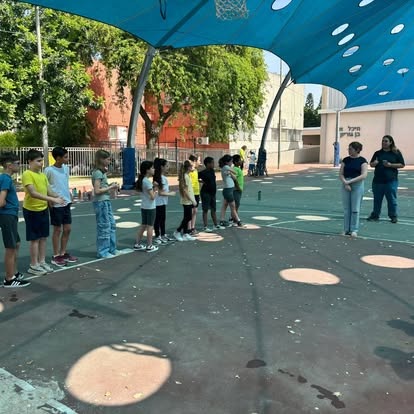 תלמידים עומדים בטור במגרש בית הספר תחת גג הצללה כחול